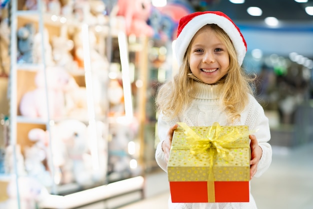 Una bambina con un cappello di natale con un regalo di capodanno in mano a una vetrina con giocattoli per bambini