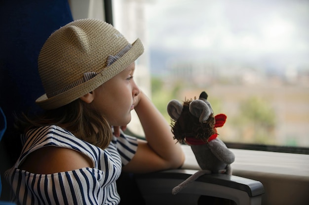 Una bambina con un cappello di paglia è seduta vicino al finestrino del treno, accanto a lei c'è il suo giocattolo