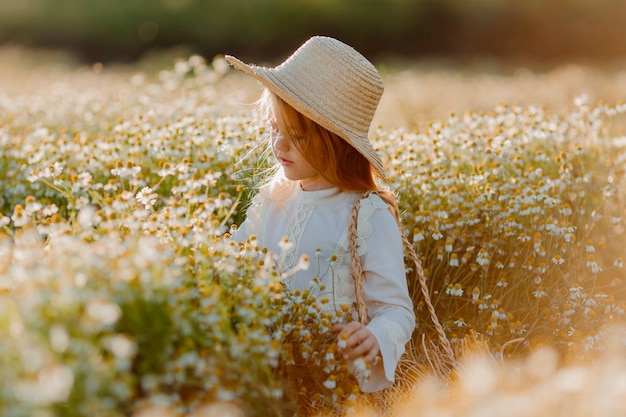 una bambina con un cappello di paglia in piedi su un campo di camomilla in una giornata di sole