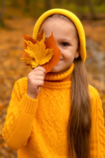 una bambina con un cappello giallo e un maglione tiene le foglie autunnali sul viso foresta autunnale urbana