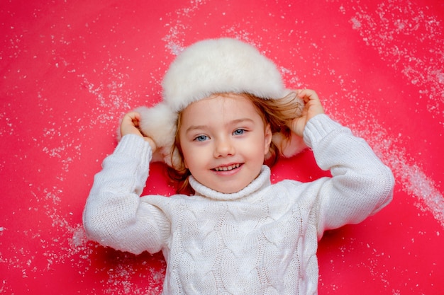 una bambina con un cappello invernale è sdraiata sul pavimento nella neve su uno sfondo rosso
