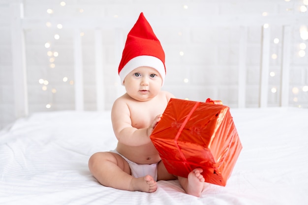 Una bambina con un cappello rosso da Babbo Natale è seduta su un letto bianco con scatole regalo su sfondo bianco
