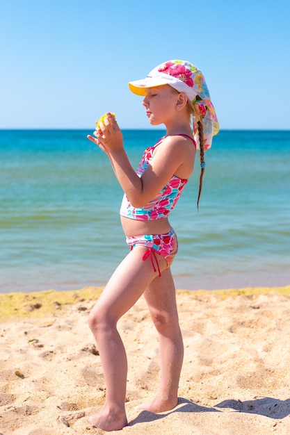 Una bambina con un codino in un cappello di Panama si trova in riva al mare e mangia mais. Vacanza al mare