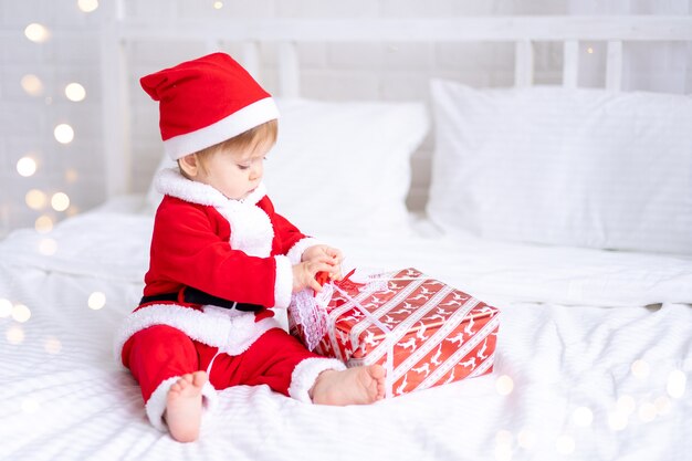 Una bambina con un costume rosso da Babbo Natale è seduta su un letto e guarda i regali di Capodanno in una scatola rossa