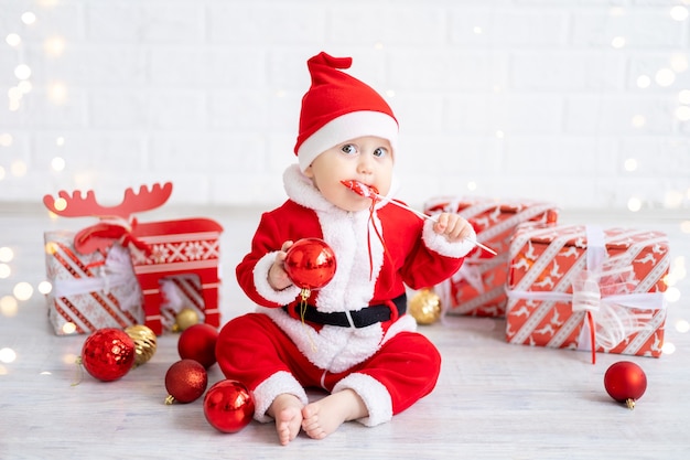 Una bambina con un costume rosso da Babbo Natale siede con scatole di regali e giocattoli di Natale su uno sfondo bianco