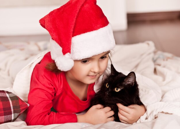 Una bambina con un divertente cappello da Babbo Natale e un pigiama natalizio giace su un plaid con un gatto nero preferito