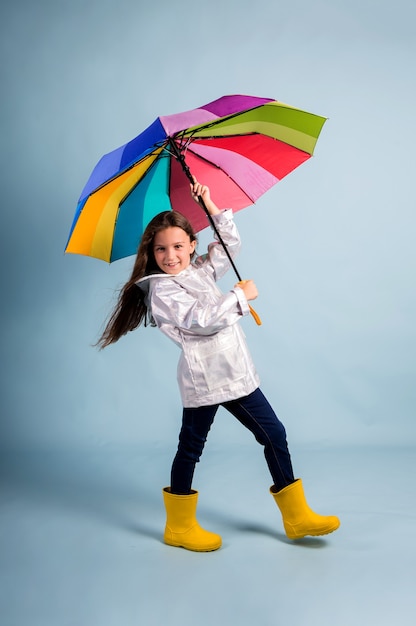 Una bambina con un impermeabile e stivali di gomma gialli sta in piedi e tiene in mano un ombrello multicolore su uno sfondo blu con un posto per il testo