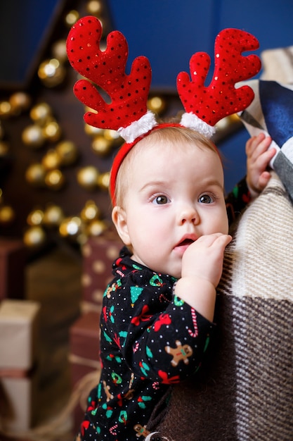 Una bambina con un maglione caldo si siede sotto un albero di Natale con giocattoli e regali con le corna in testa. Infanzia felice. Atmosfera di festa di Capodanno