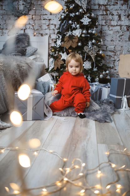 Una bambina con un maglione rosso caldo si siede sotto un albero di Natale con giocattoli e regali. Infanzia felice. Atmosfera di festa di Capodanno