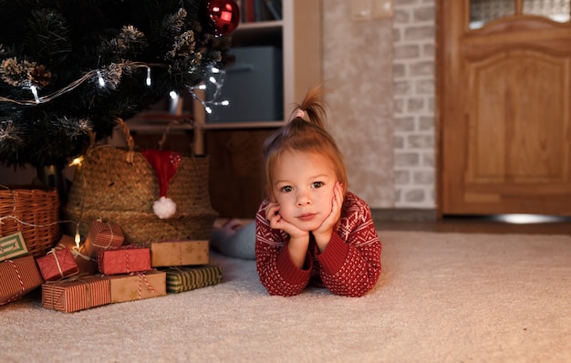 Una bambina con un maglione rosso si trova sotto l'albero tra i regali di Natale.