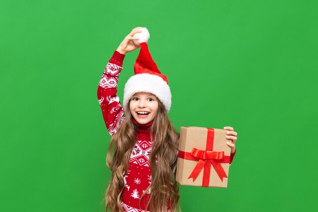 Una bambina con un regalo in mano si tiene per il cappello di Babbo Natale e sorride ampiamente su uno sfondo verde isolato Regali di Natale per bambini