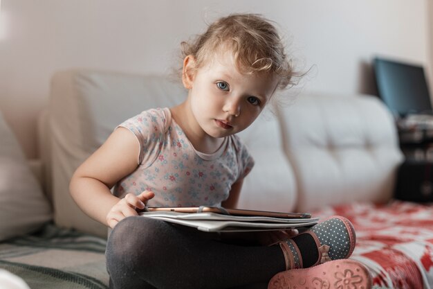 Una bambina con un tablet si siede sul divano, studia a casa