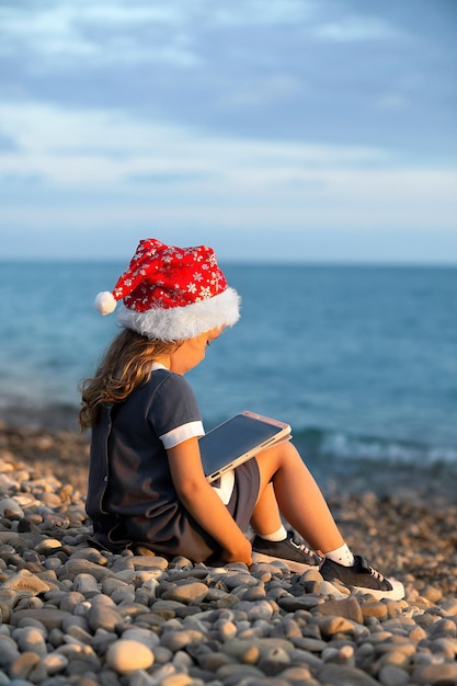 Una bambina con un vestito blu e un cappello rosso di Babbo Natale si siede in riva al mare