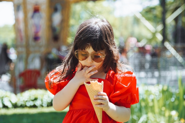 Una bambina con un vestito rosso e un cappello di paglia mangia il gelato in un cono nel parco