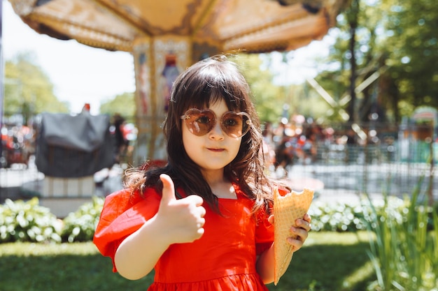 Una bambina con un vestito rosso e un cappello di paglia mangia il gelato in un cono nel parco