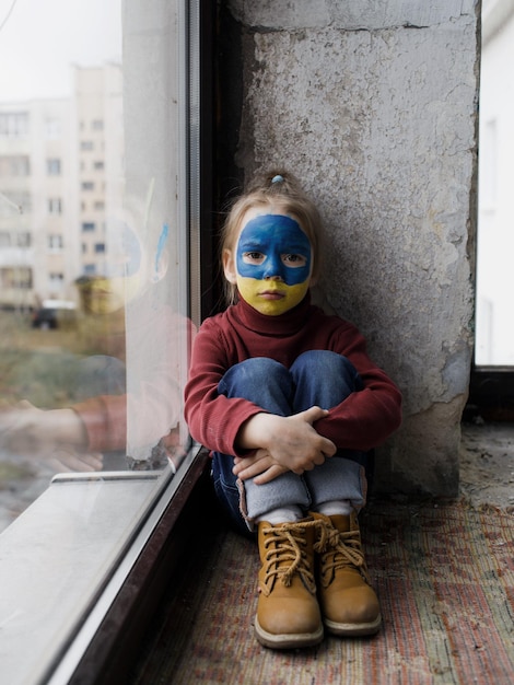 Una bambina con una bandiera ucraina dipinta sul viso è seduta coperta da una coperta vicino alla finestra I bambini e la guerra in Ucraina