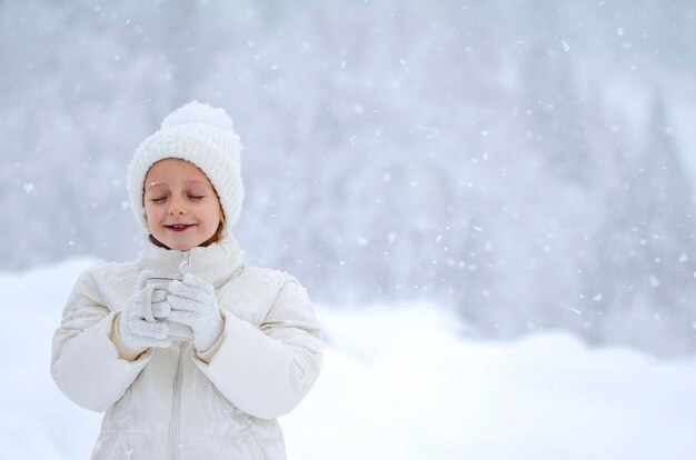 Una bambina con una giacca bianca e un cappello bianco tiene in mano una tazza di tè durante una nevicata e cattura i fiocchi di neve con la lingua sullo sfondo di bellissimi cumuli di neve e foreste