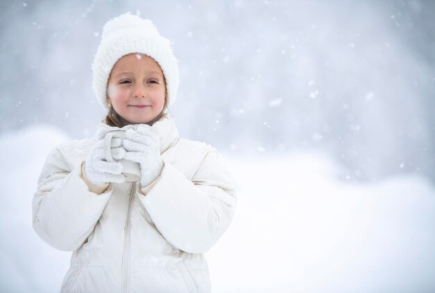 Una bambina con una giacca bianca e un cappello bianco tiene in mano una tazza di tè durante una nevicata sullo sfondo di bellissimi cumuli di neve e foreste