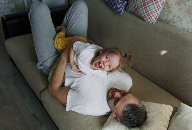 Una bambina con una maglietta bianca si sta divertendo a giocare con suo padre sul divano, divertendosi a vicenda. Si guardano l'un l'altro.