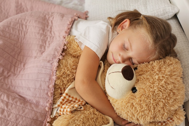 una bambina con una maglietta bionda dorme a casa sotto una coperta rosa abbracciando un orsacchiotto