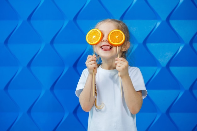 Una bambina con una maglietta sorride e tiene due metà di frutta arancione davanti ai suoi occhi su uno sfondo blu. Concept: la lotta all'avitomnosi primaverile.