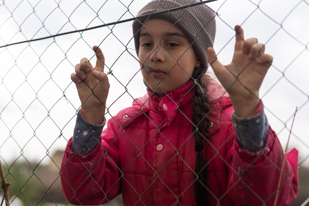 Una bambina con uno sguardo triste dietro una recinzione metallica. Problema sociale dei rifugiati e dei migranti forzati