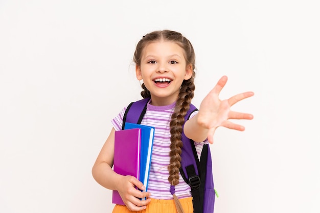 Una bambina con uno zaino e libri tira la mano in avanti su uno sfondo bianco isolato Il bambino ti invita ai corsi