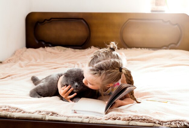 Una bambina dai capelli biondi giace su un letto a casa con un gatto che legge un libro. Resta a casa. Istruzione a casa per i bambini.