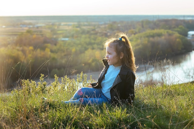 una bambina dai capelli mossi siede su un'alta sponda del fiume al tramonto