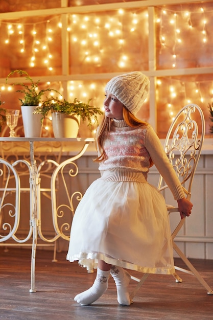 Una bambina dai capelli rossi brillante con un cappello bianco e un bel vestito è seduta in un caffè autunnale. Ragazza dai capelli rossi con grandi occhi azzurri e un bel maglione incontra la primavera