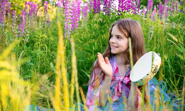 Una bambina di 10 anni con i capelli lunghi siede in una radura con fiori di lupino e gioca con un tamburello