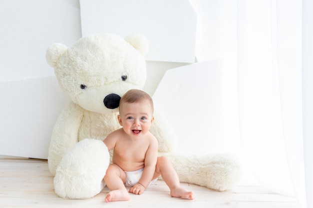 Una bambina di 6 mesi è seduta con un grande orso molle in un luminoso appartamento con i pannolini