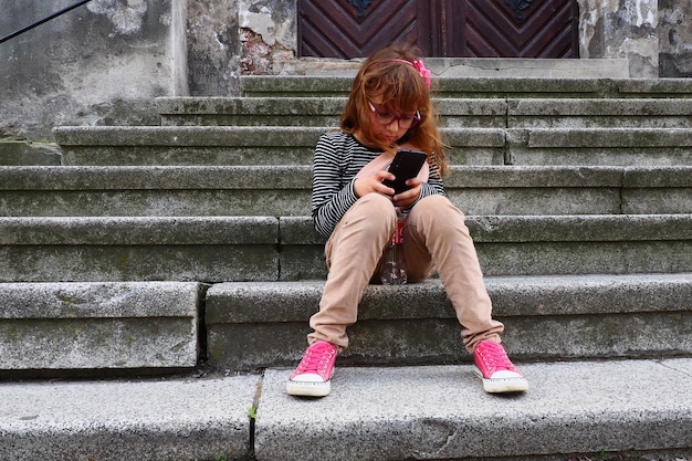 Una bambina di 8 anni con gli occhiali siede sulle scale di una chiesa a Petrovaradin guarda e usa un telefono o uno smartphone Vita cittadina e comunicazioni Ritmo moderno Dipendenza da telefono e internet