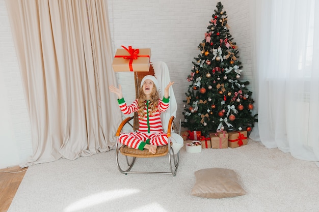Una bambina di 9 anni con in mano un regalo di Babbo Natale e seduta su una sedia a dondolo con ghirlande sullo sfondo. Periodo natalizio. Buon anno!