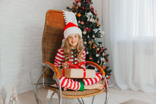 Una bambina di 9 anni con in mano un regalo di Babbo Natale e seduta su una sedia a dondolo con ghirlande sullo sfondo. Periodo natalizio. Buon anno!