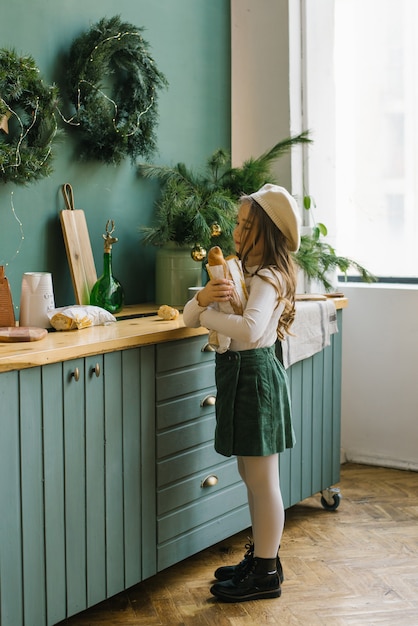 Una bambina di cinque anni in eleganti abiti di fiori bianchi e verdi tiene in mano un sacchetto di carta con una baguette, in piedi vicino alla cucina, decorata per Natale