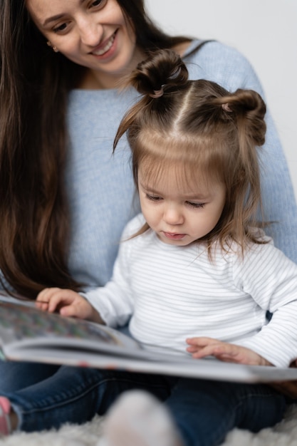 Una bambina di due anni è seduta con la madre in grembo. Resta a casa. La mamma con la figlia legge un grande libro di carta. Racconti per bambini interessanti