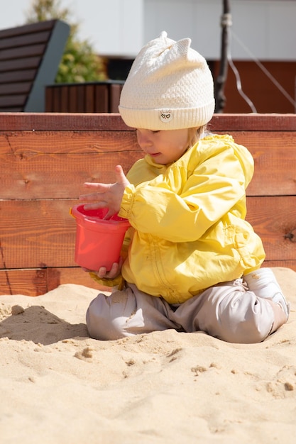 Una bambina di due anni è seduta nella sandbox e gioca con un secchio e della sabbia