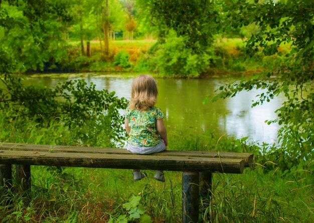 Una bambina di due anni seduta da sola sulla panchina e guardando il lago