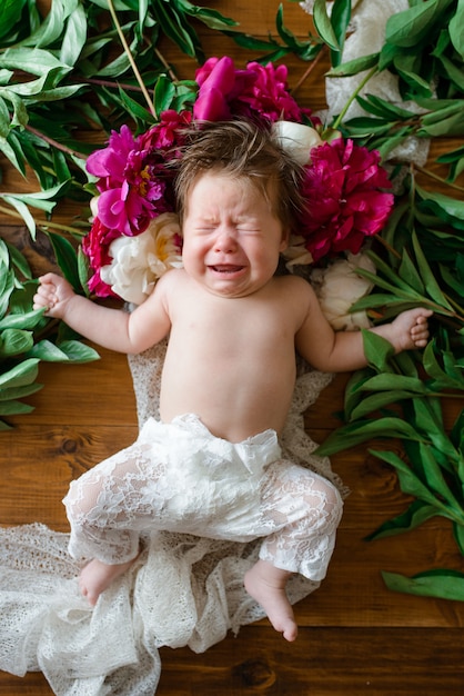 Una bambina di due mesi giace su un tavolo di legno con peonie e pianti.