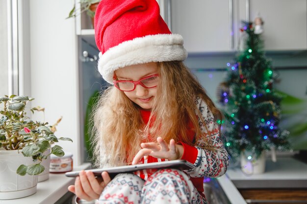 Una bambina di quattro anni con un cappello da Babbo Natale e occhiali scrive una lettera sul taccuino a Babbo Natale mentre è seduta sul tavolo della cucina