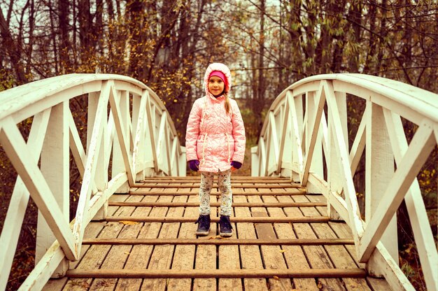 Una bambina di sei anni felice in abiti autunnali si trova sul ponte di legno a schiena d'asino nel parco autunnale