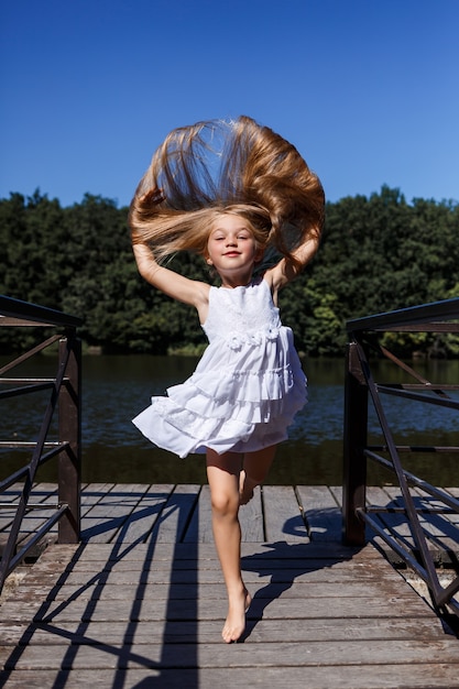 Una bambina di sei anni indossa un abito bianco. Bellissimi capelli lunghi. Cammina vicino al fiume sullo sfondo della foresta