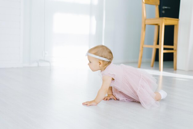 Una bambina di un anno con un vestito rosa striscia sul pavimento della stanza dei bambini