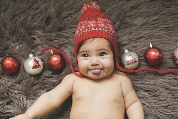 Una bambina di un anno giace su un copriletto, con un cappello rosso e gioca con i giocattoli di Capodanno