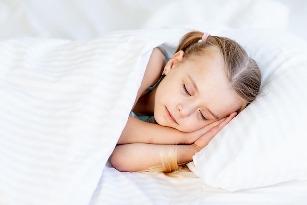 Una bambina dorme su un letto a casa su un letto di cotone bianco e sorride dolcemente nel sonno con le mani giunte sotto le guance