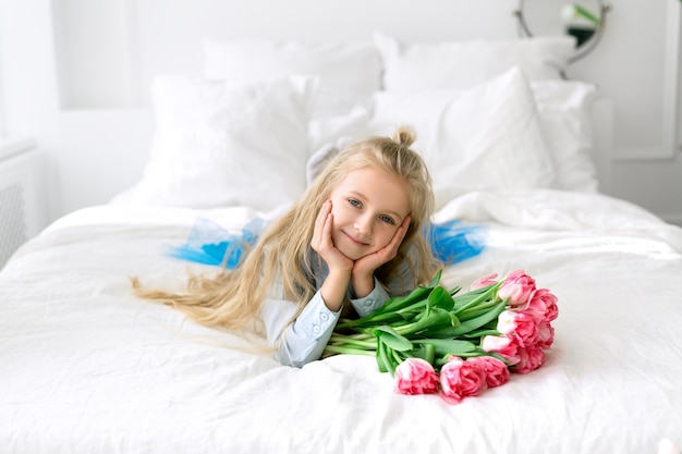 Una bambina è sdraiata su un letto bianco con un mazzo di tulipani rosa