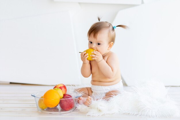 Una bambina è seduta in una stanza luminosa con un piatto di frutta e sta mangiando una pera