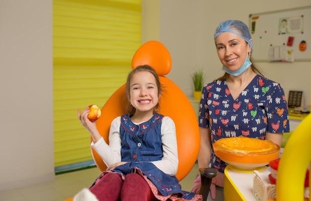 Una bambina è seduta su una poltrona odontoiatrica arancione sorridente con una mela rossa in mano Pediatrica