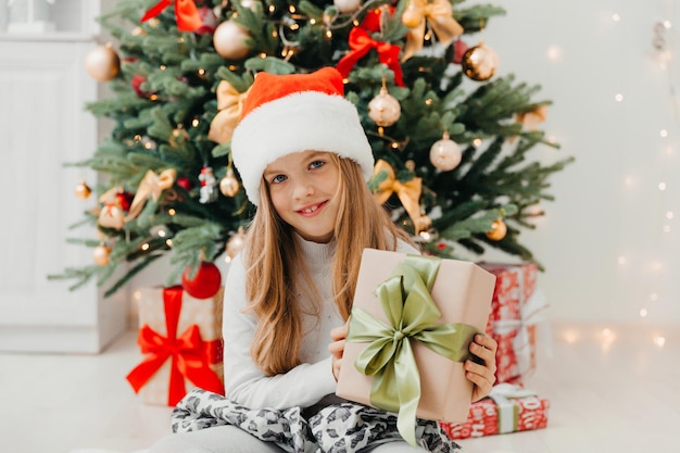 Una bambina è seduta vicino all'albero di Natale con un regalo di Natale. Buon Natale.